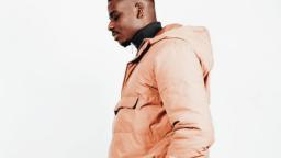 Image of Dami Sule. He is standing to the side, and wearing a beige hooded jacket over a black polo neck jumper.